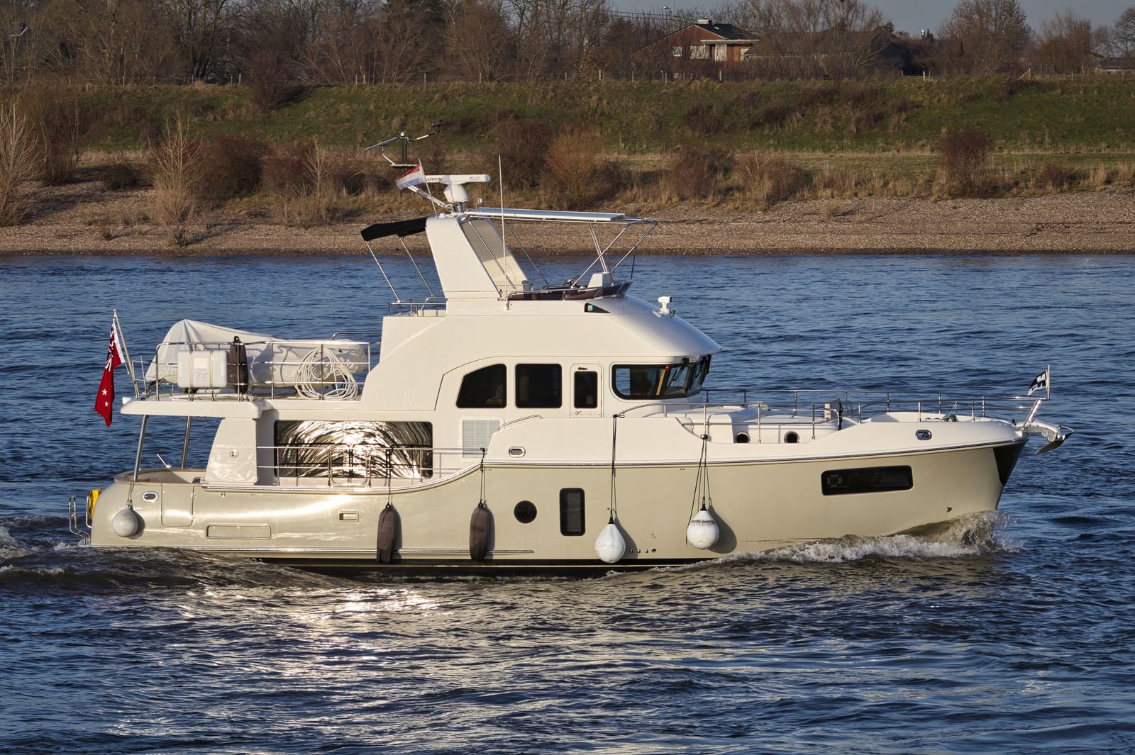 Trawler yacht underway on a river, running at displacement speed with a dinghy on the upper deck